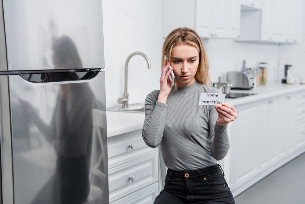 serious young woman holding card with lettering home inspection and talking on smartphone near